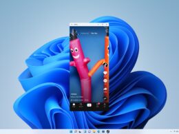 Úzke zvislé okno aplikácie TikTok s prehrávaným videom dvoch nafukovacích tancujúcich panákov stojí uprostred plochy Windows 11 s modrou vlnitou tapetou a vycentrovaným panelom úloh.