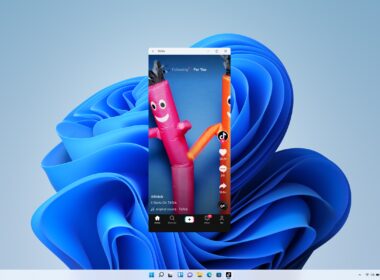 Úzke zvislé okno aplikácie TikTok s prehrávaným videom dvoch nafukovacích tancujúcich panákov stojí uprostred plochy Windows 11 s modrou vlnitou tapetou a vycentrovaným panelom úloh.