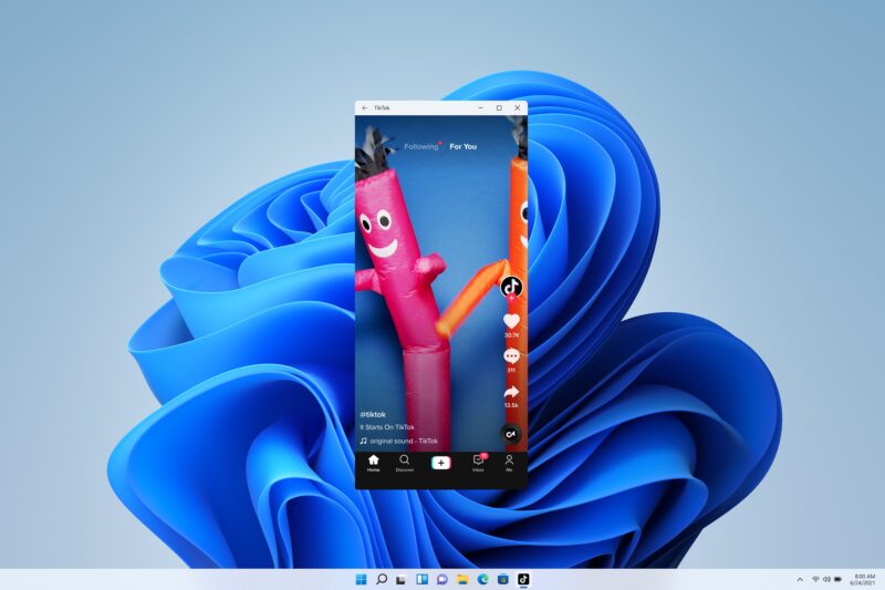 Úzke zvislé okno aplikácie TikTok s prehrávaným videom dvoch nafukovacích tancujúcich panákov stojí uprostred plochy Windows 11 s modrou vlnitou tapetou a vycentrovaným panelom úloh.