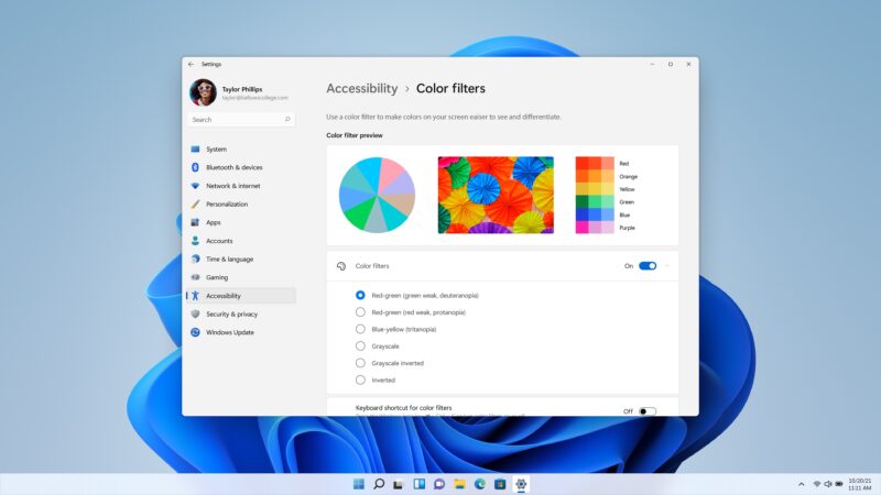 Okno Nastavení Windows 11 otvorené na stránke Prístupnosť – Farebné filtre s ukážkou farebného kolesa, fotografiou farebných dáždnikov a paletou farieb, pod ňou zapnutý prepínač a zoznam filtrov s vybranou možnosťou pre deuteranopiu, na pozadí modrá tapeta Bloom.