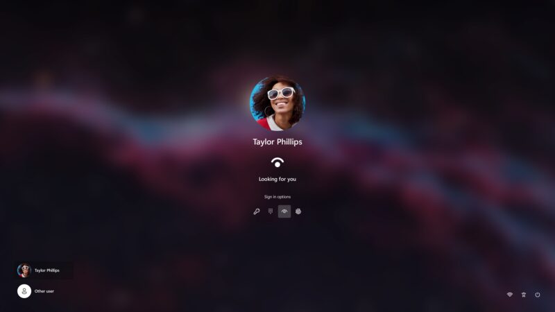 Prihlasovacia obrazovka Windows 11 na tmavom rozostrenom pozadí, v strede kruhový profilový obrázok usmievajúcej sa ženy v bielych okuliaroch, pod ním meno Taylor Phillips, ikona snímania tváre a text „Looking for you“, nižšie možnosti prihlásenia – kľúč, PIN, tvár a odtlačok prsta; vľavo dole zoznam používateľov, vpravo dole Wi-Fi, prístupnosť a vypnutie.