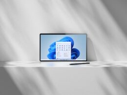 Tablet opretý o svetlú stenu, po ktorej padajú tiene žalúzií, má na obrazovke modrú tapetu Windows 11 s otvorenou ponukou Štart a vedľa neho leží na doske tmavé dotykové pero.