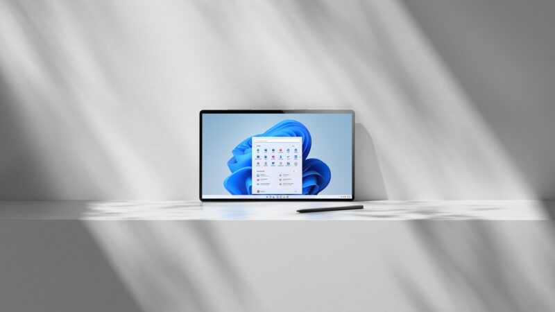 Tablet opretý o svetlú stenu, po ktorej padajú tiene žalúzií, má na obrazovke modrú tapetu Windows 11 s otvorenou ponukou Štart a vedľa neho leží na doske tmavé dotykové pero.