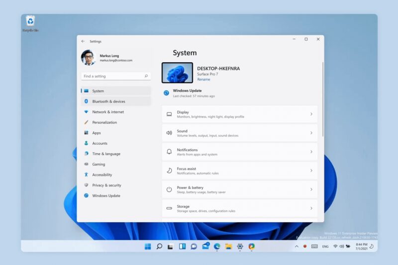 Okno Nastavení Windows 11 otvorené na stránke Systém, s bočnou ponukou položiek ako Bluetooth a zariadenia, Personalizácia či Windows Update a s dlaždicami Obrazovka, Zvuk a Oznámenia; pod oknom je panel úloh so zarovnanými ikonami v strede.